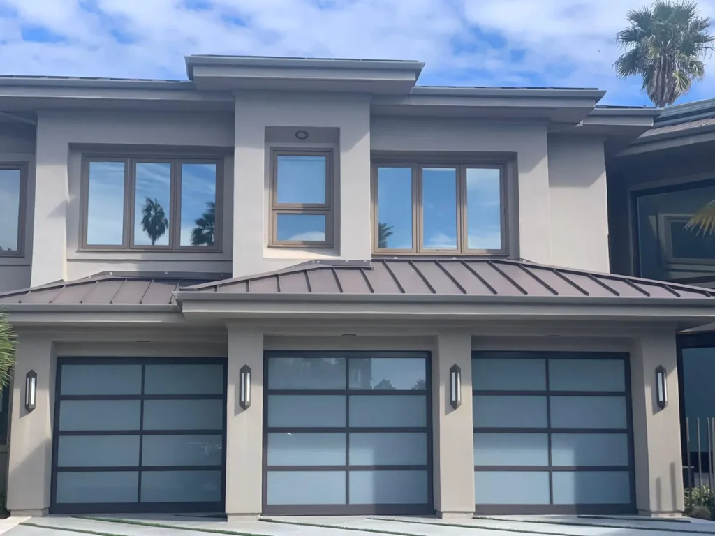 A modern two-story grey stucco house with a dark metal roof. The front features large windows and two contemporary sectional garage doors made of blue-tinted glass and thin metal frames.